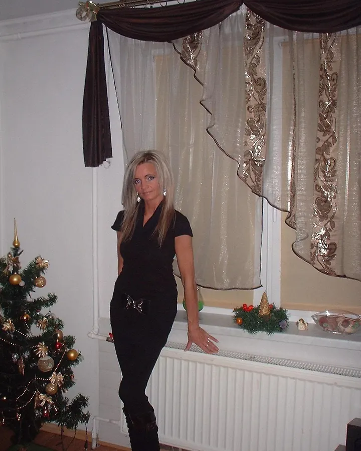 Femme aux cheveux blonds, vêtue d'une robe noire ajustée, debout dans une pièce décorée de Noël, près d'un arbre et d'une fenêtre avec rideaux dorés.