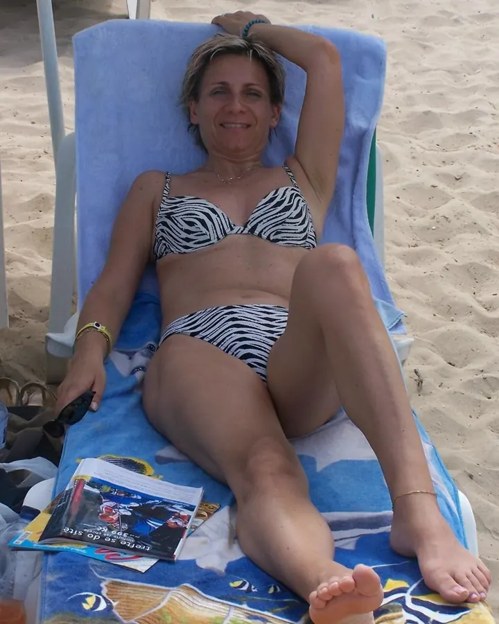Femme aux cheveux blonds courts, portant un haut blanc sans manches et une jupe noire, assise sur un transat sur une plage, avec un maillot de bain zébré noir et blanc.