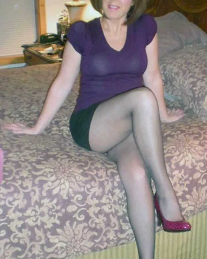 Femme aux cheveux blonds courts, en robe noire à sequins, assise sur un canapé beige, souriant à la caméra.