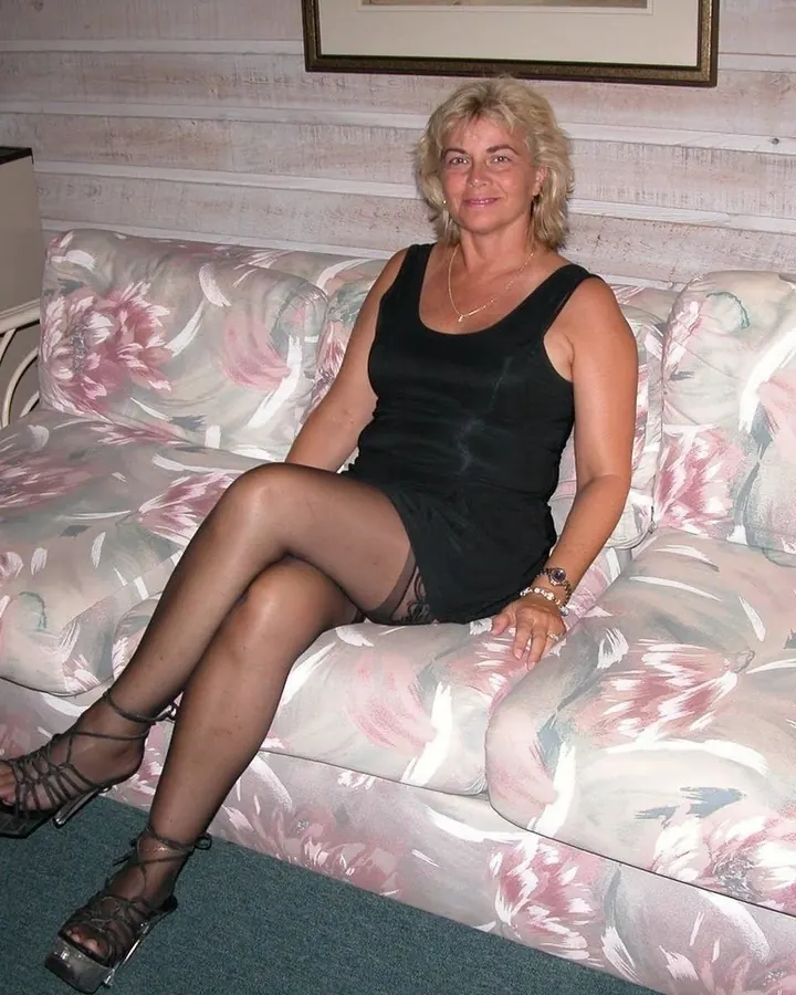 Femme aux cheveux blonds, vêtue d'une tenue noire, posant sur un canapé beige dans un salon élégant.