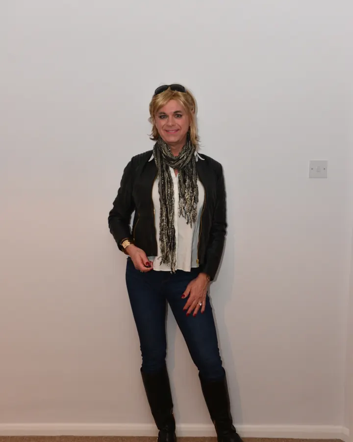 Femme blonde en veste en cuir noir, jeans, bottes hautes, foulard, souriant devant un mur blanc.