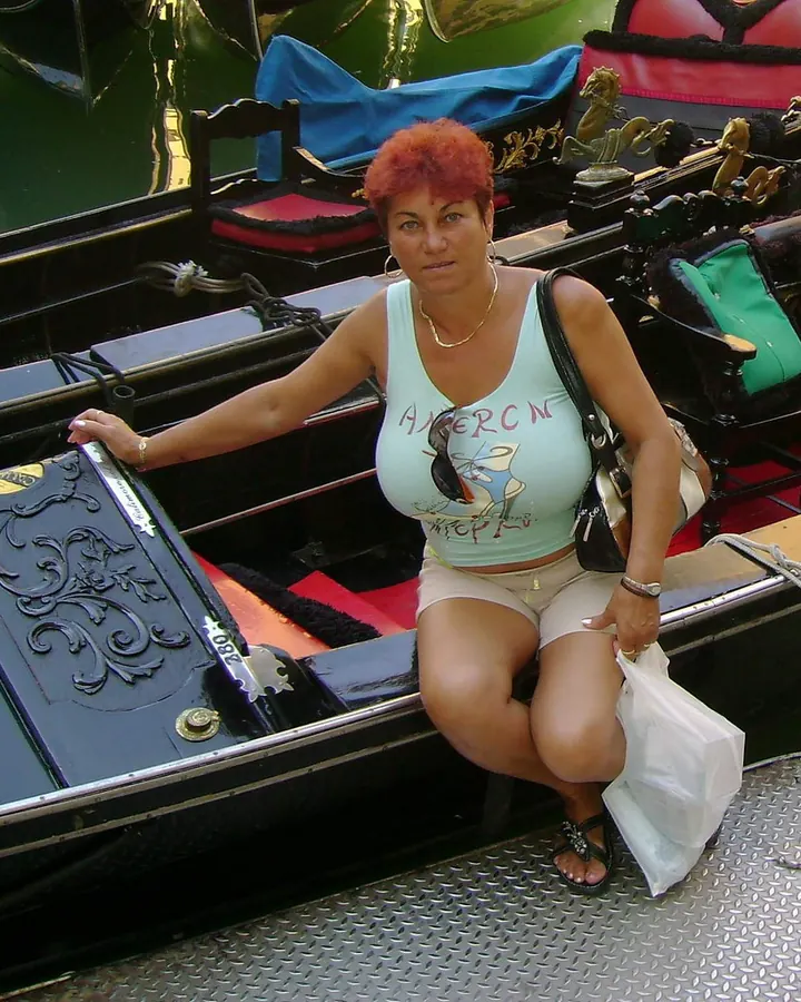 Femme assise sur un bateau décoratif, en haut vert clair et short beige, avec des lunettes de soleil et des cheveux roux courts.