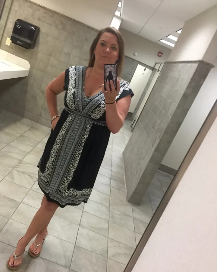 Femme prenant un selfie dans un miroir, en robe noire et blanche, dans un WC public.